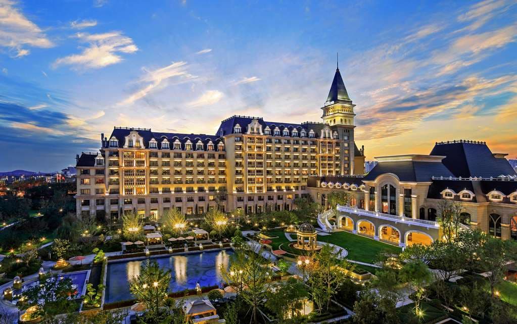 A twilight view of the castle-like Hilton Qingdao Golden Beach resort illuminated against a dark blue sky, with the outdoor pool glowing in the foreground.