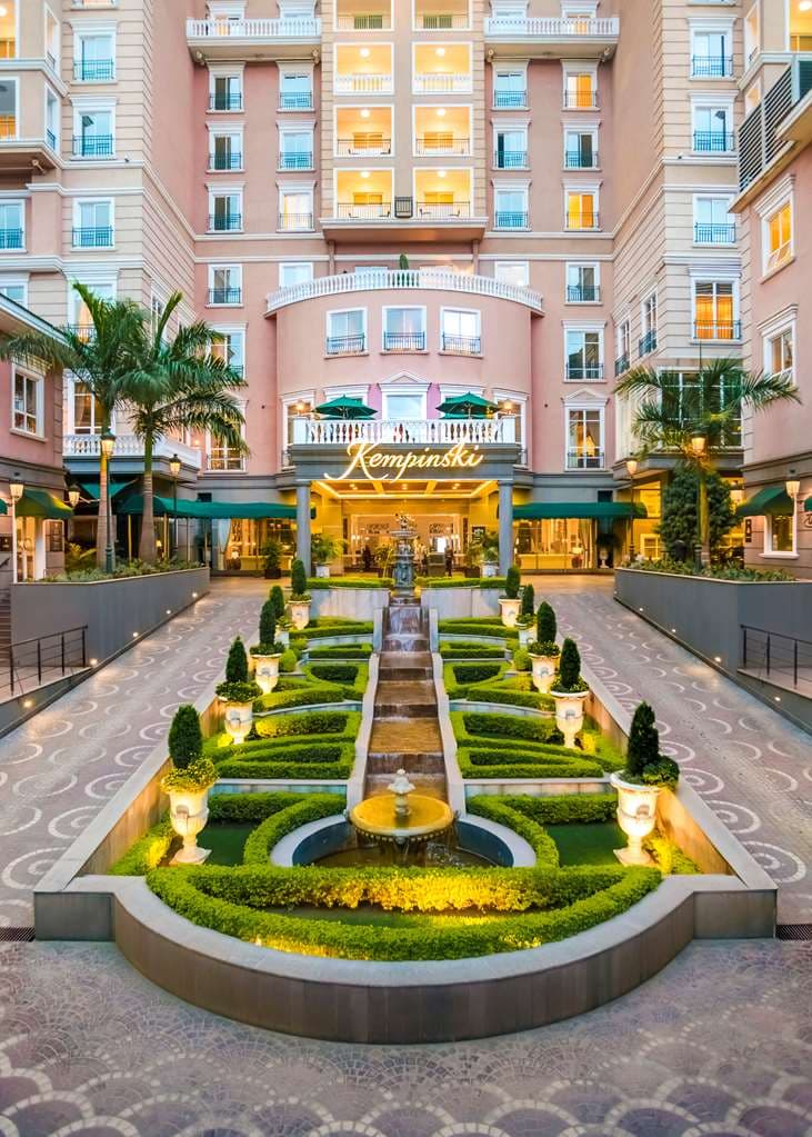 The pink-hued exterior of Villa Rosa Kempinski Nairobi featuring the outdoor heated pool and sun loungers against a backdrop of lush green trees.