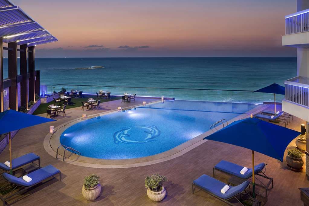A dramatic golden-hour view from the Hilton Alexandria Corniche rooftop infinity pool, overlooking the curved Mediterranean coastline and the bustling El Geish Road.