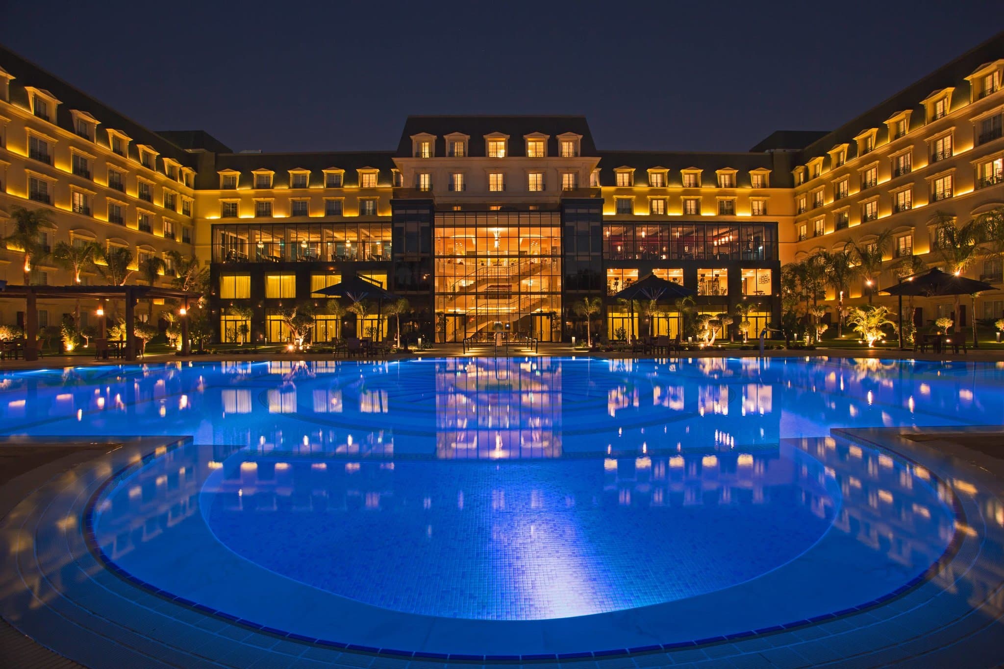 The European-style central pool deck of the Renaissance Cairo Mirage City Hotel at twilight, framed by palm trees and illuminated facade arches.