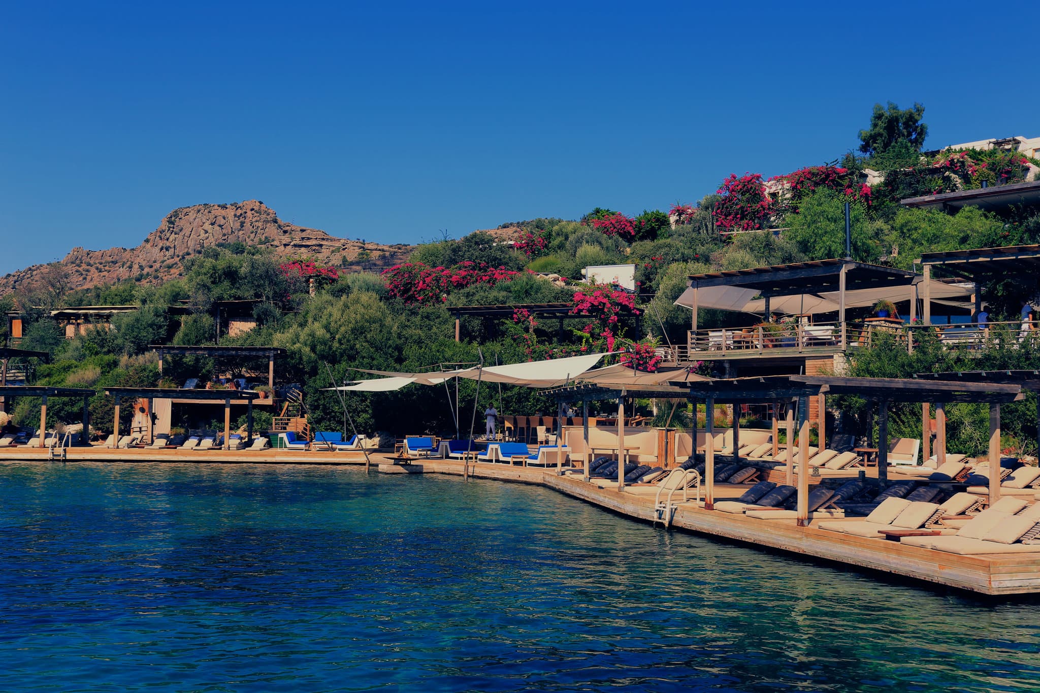 The iconic wooden sun decks of Maçakızı Hotel lining the turquoise Aegean Sea, dotted with white umbrellas and chic sunbathers.