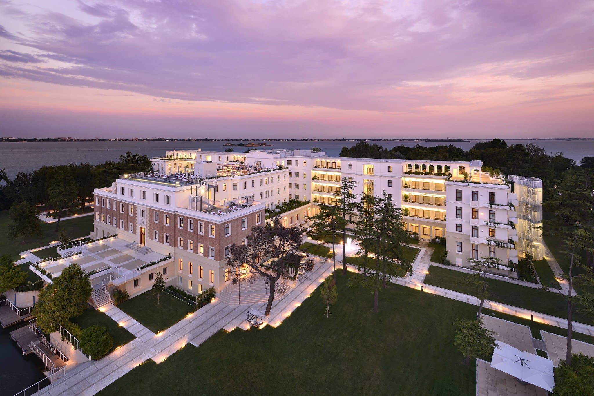 A sleek, white modernist building on a private green island in the Venetian lagoon, framed by olive trees with the Venice skyline in the distance.