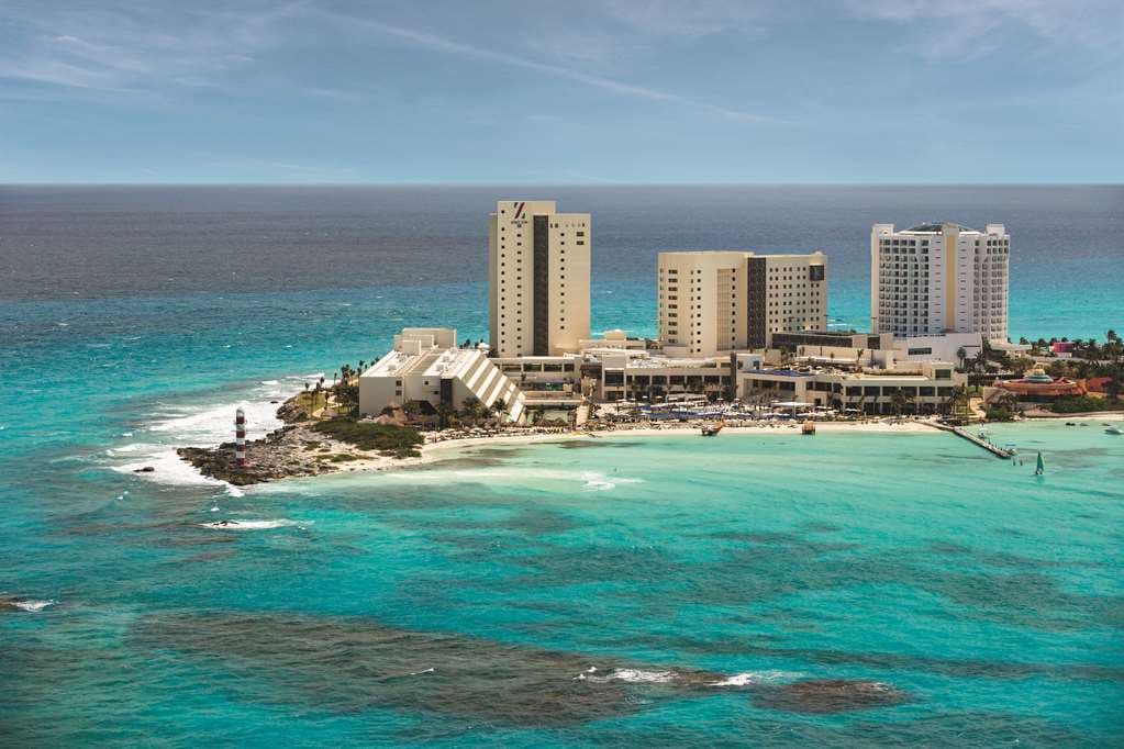 Aerial view of Hyatt Ziva Cancun on the Punta Cancun peninsula, surrounded by turquoise waters on three sides with the red and white lighthouse in the background.