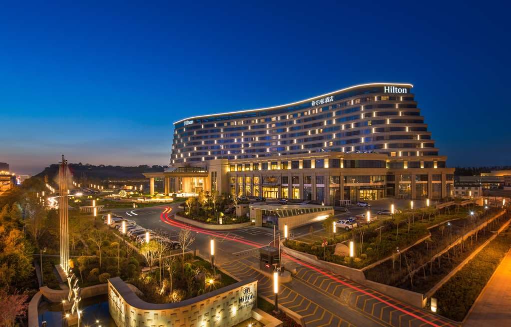 A dramatic low-angle shot of the Hilton Urumqi's glass facade reflecting the golden hues of the Hongguang Hill Buddha statue.