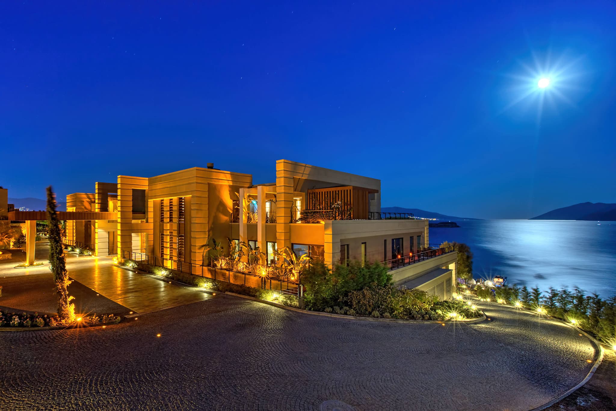 View of the Caresse Bodrum infinity pool blending into the turquoise Aegean Sea, with the Buddha-Bar Beach deck and lounging guests in the background.