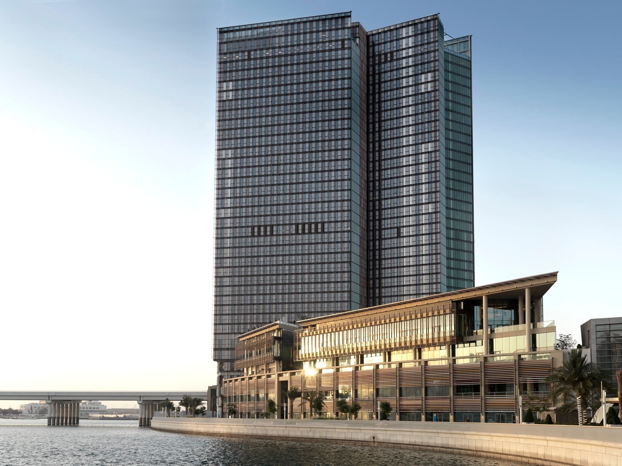 A low-angle shot of the Four Seasons Abu Dhabi exterior featuring vertical terracotta baguettes against a blue sky, adjacent to the water.