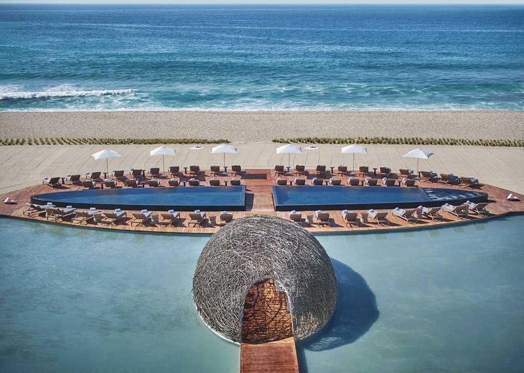The NIDO restaurant at Viceroy Los Cabos, a nest-like wooden structure illuminated at night floating on mirror pools surrounded by white concrete buildings.