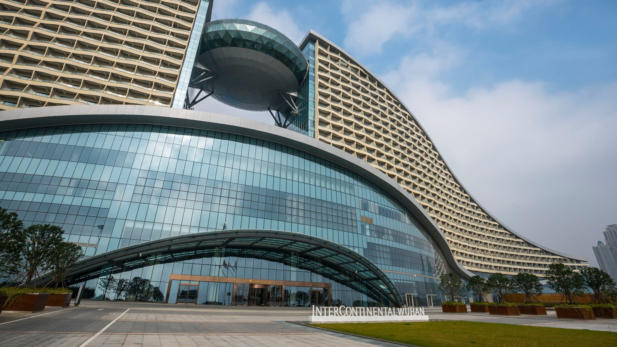 A low-angle view of the InterContinental Wuhan's futuristic, geometric glass atrium glowing golden at dusk against a deep blue sky.