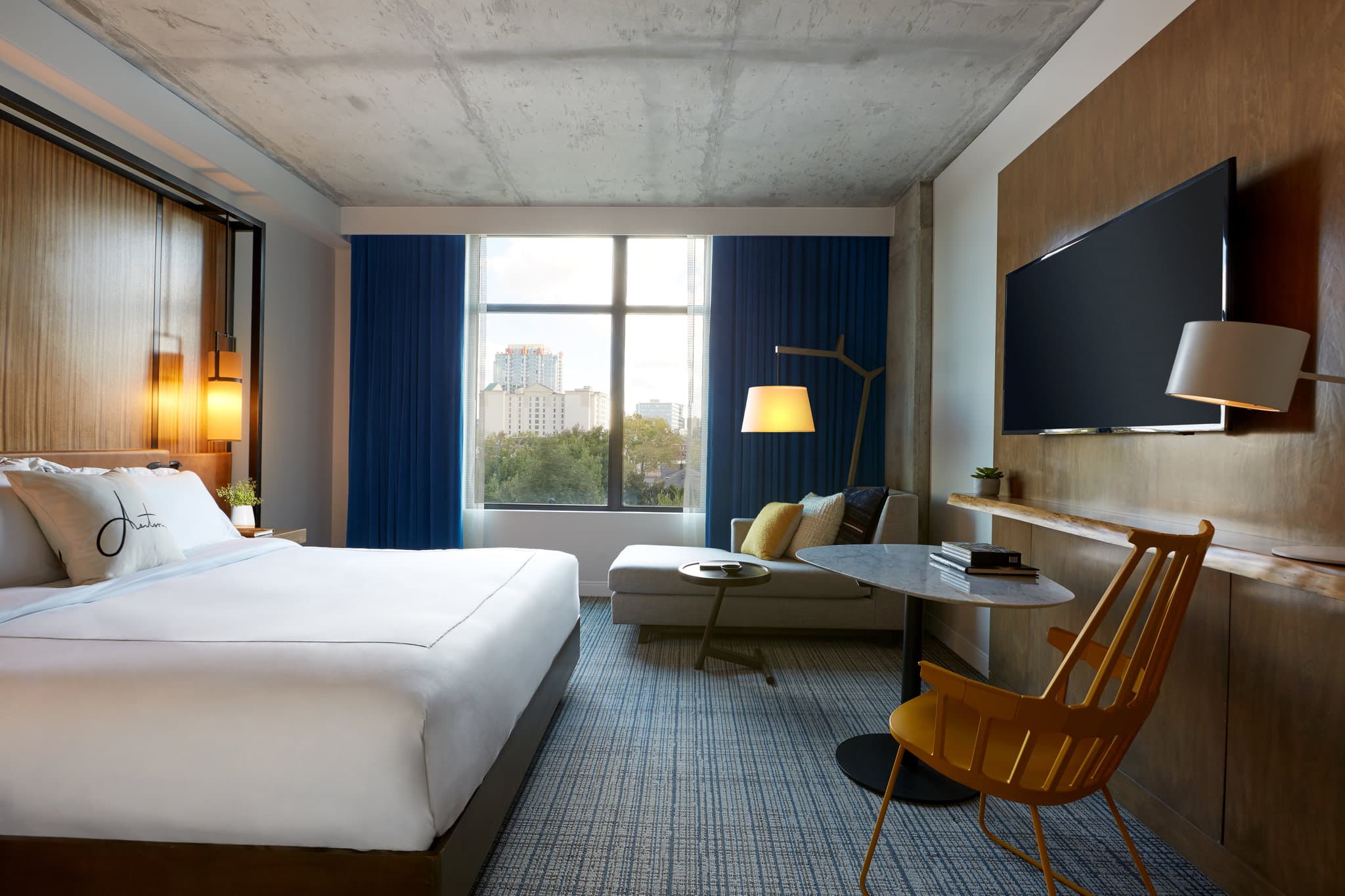 A moody, industrial-chic hotel room at Kimpton Aertson Nashville featuring exposed concrete ceilings, a leather headboard, and large windows overlooking Vanderbilt University.