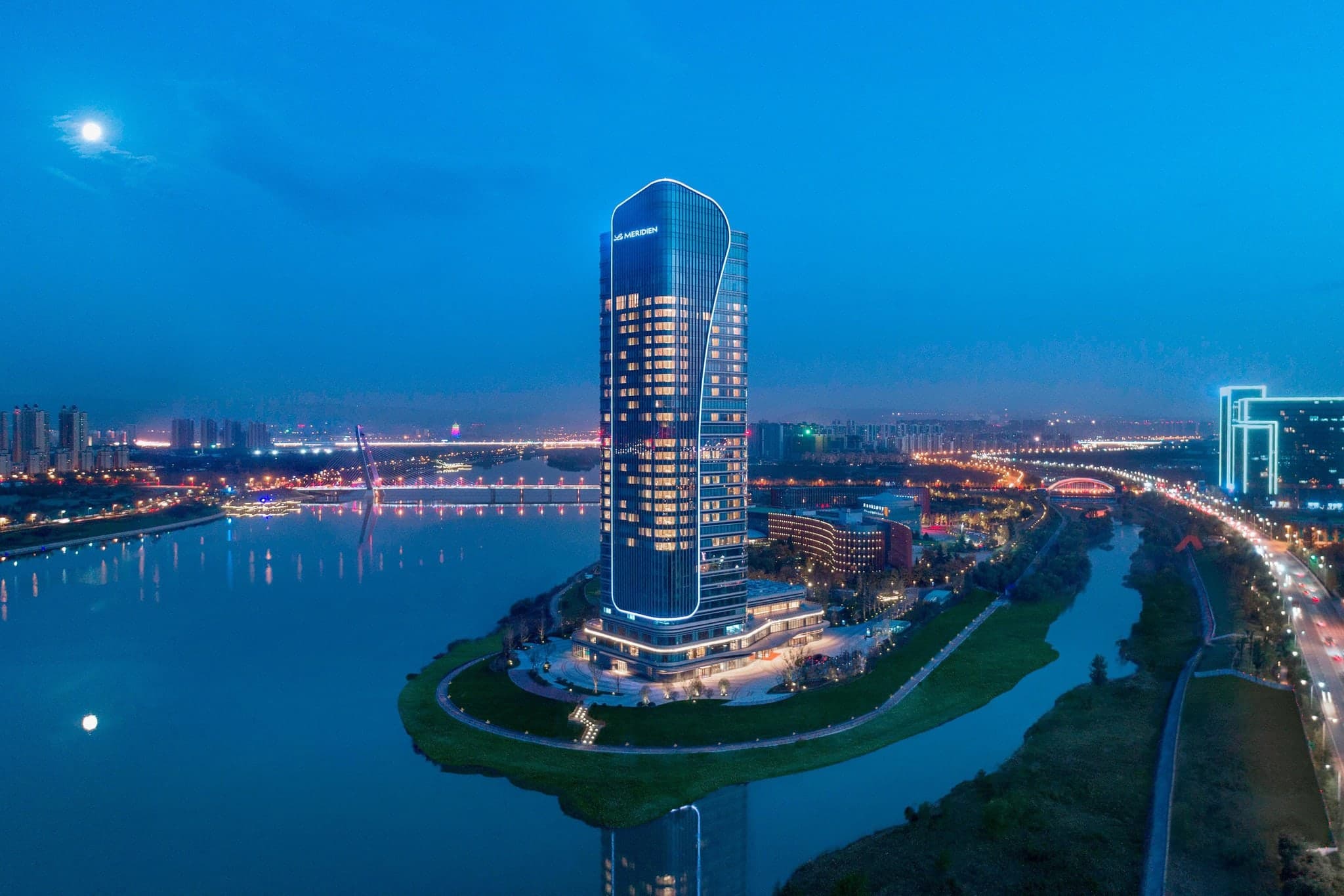 A woman relaxing in a freestanding bathtub in a Le Méridien corner suite, overlooking the winding Ba River and Xi'an skyline through floor-to-ceiling windows.