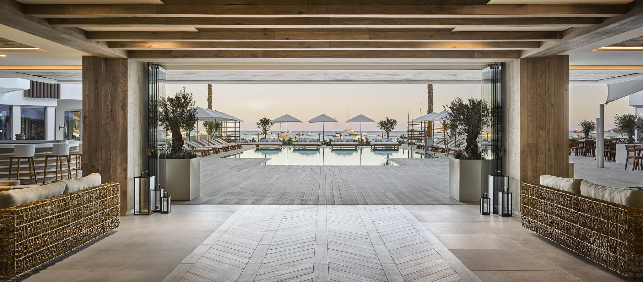 A chic wooden walkway leading to the Nobu Ibiza Bay pool area, lined with white umbrellas, palm trees, and woven rattan lamps against a bright blue sky.