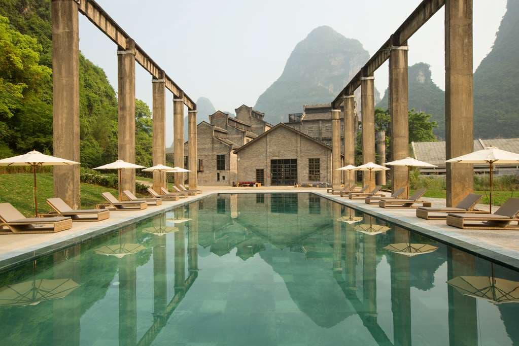 The Yangshuo Sugar House swimming pool, built on a former loading dock, framed by industrial trusses and dramatic limestone karst mountains at dusk.