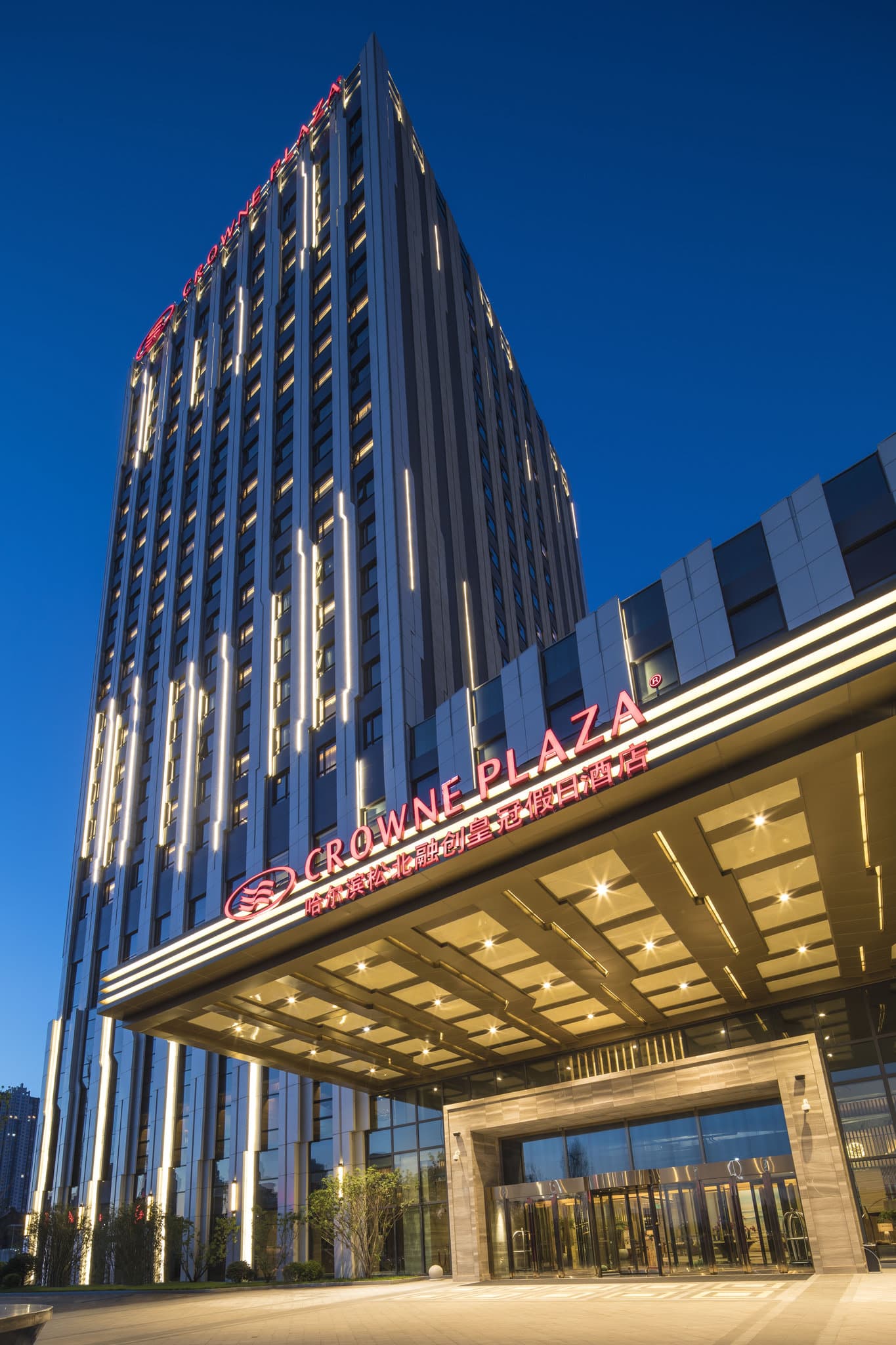 A high-angle view of the Crowne Plaza Harbin Songbei lobby featuring a massive crystal chandelier and warm, golden lighting contrasting with the winter exterior.