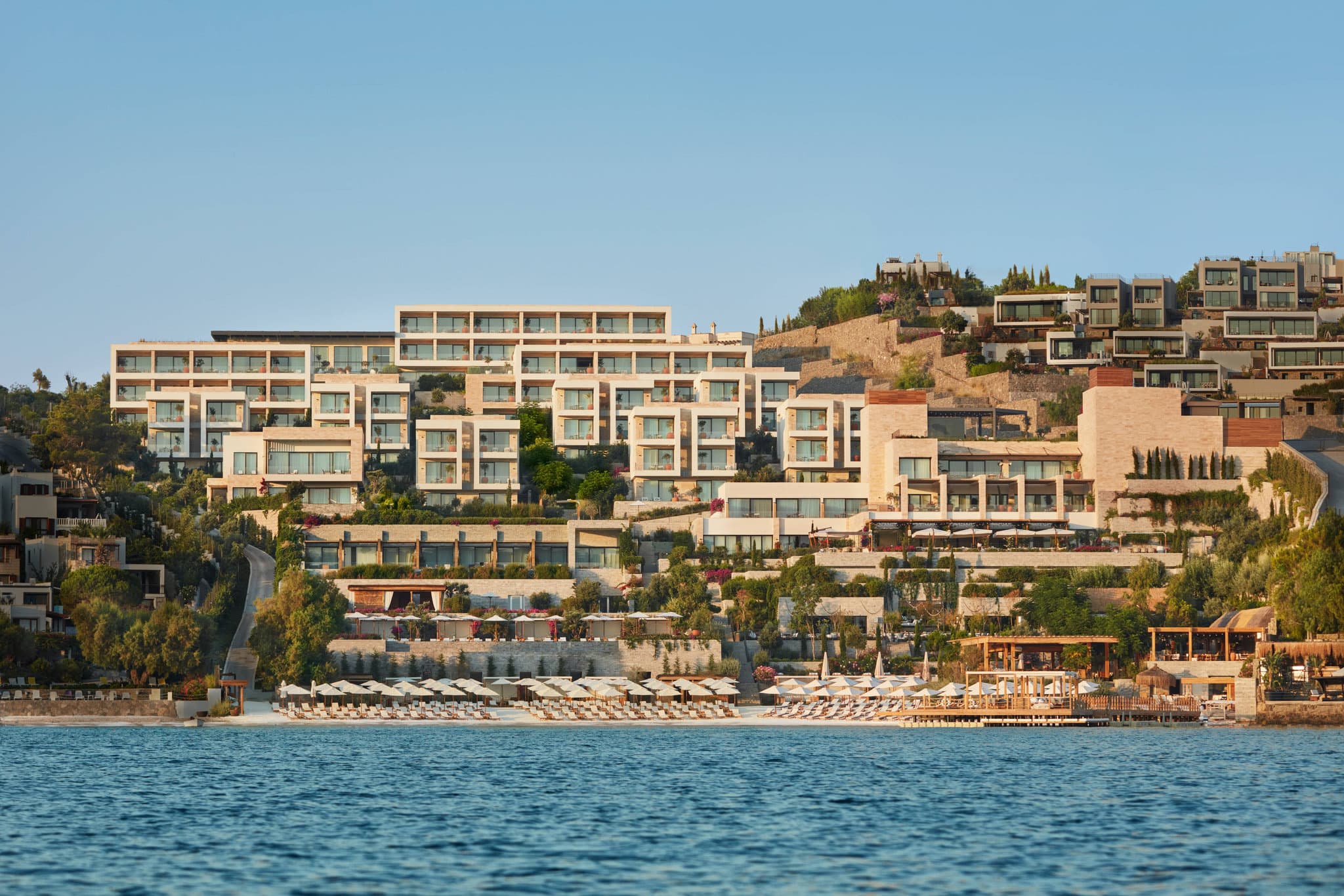 The Bodrum EDITION infinity pool merging with the turquoise Aegean Sea, framed by white umbrellas and minimalist decking.