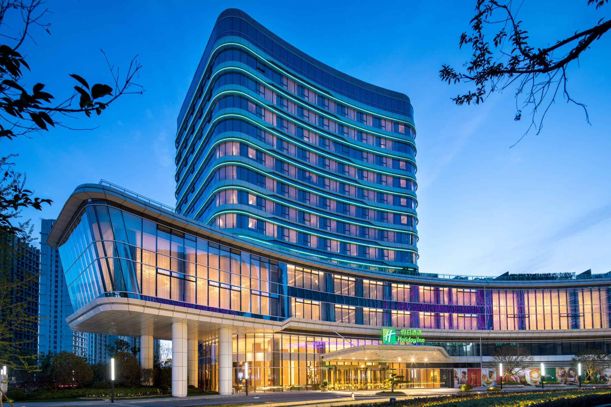 A sleek, modern hotel room at Holiday Inn Zhengzhou Riverside featuring floor-to-ceiling windows overlooking the city skyline at dusk.