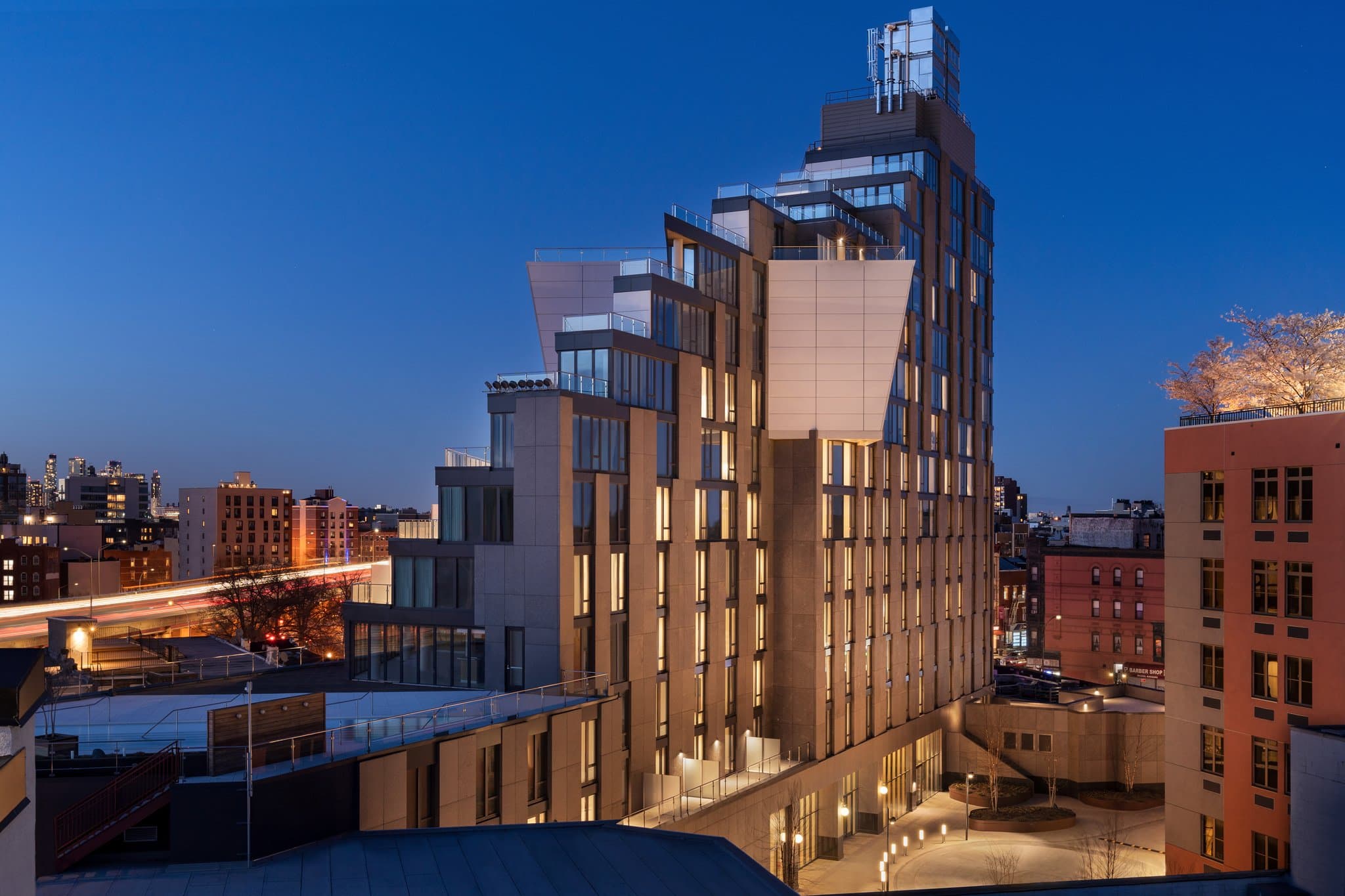 A sleek, modern hotel room at Hotel Indigo Williamsburg featuring industrial design elements, a king bed with gray linens, and a large window overlooking the Brooklyn cityscape.