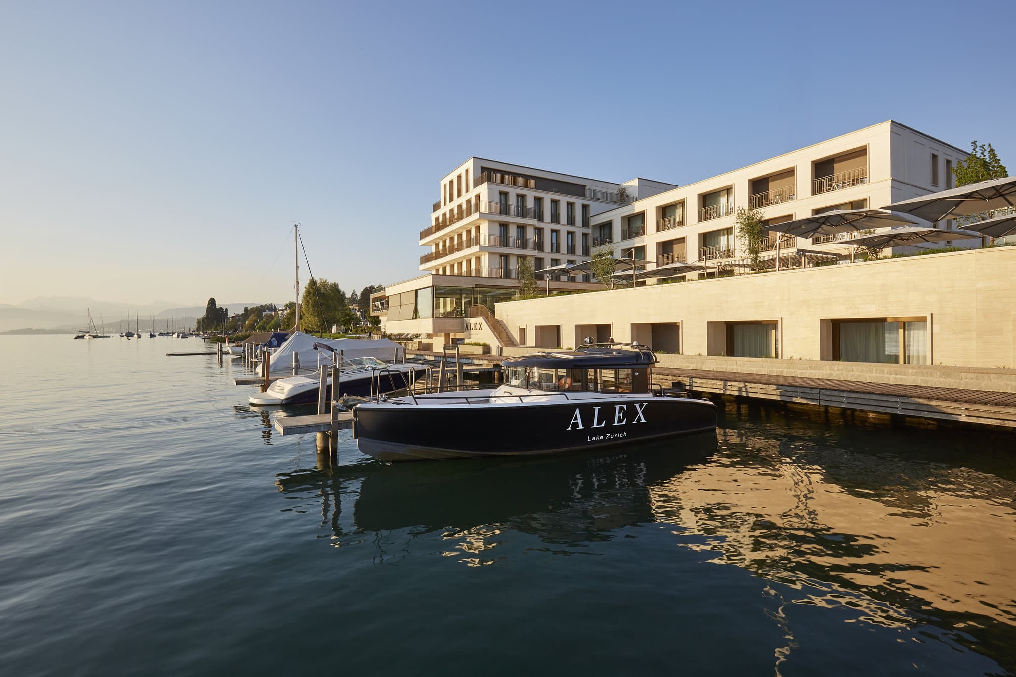 A sleek, modern terrace suite at Alex Lake Zurich overlooking the water at dusk, with a private motorboat docked below.