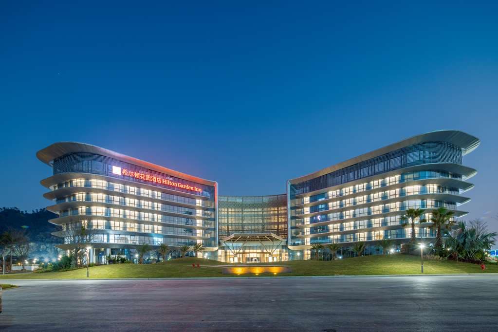 Exterior of Hilton Garden Inn Zhuhai Hengqin Sumlodol Park featuring its distinct seagull-shaped roof against a lush mountain and lake backdrop.