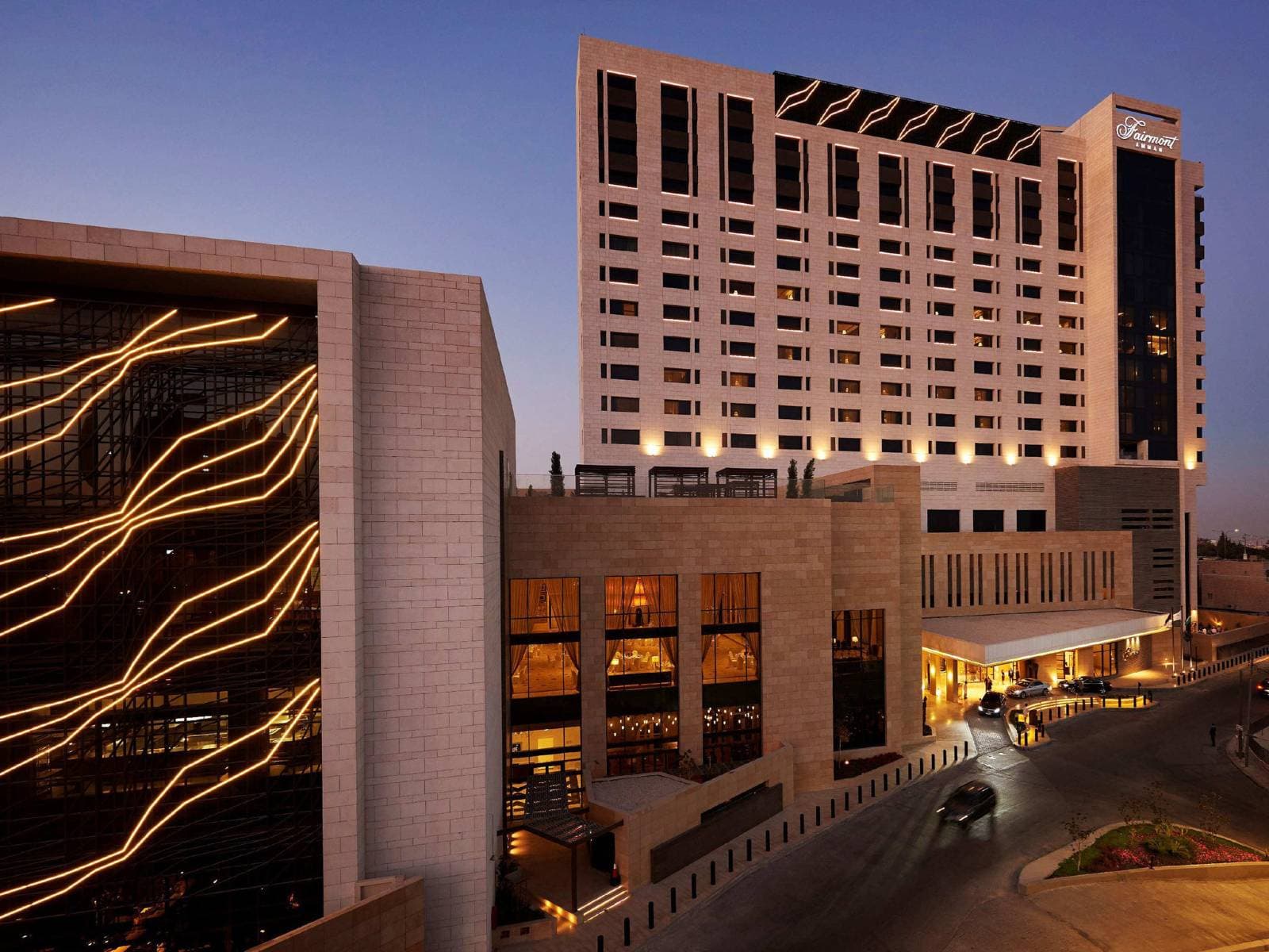 The illuminated limestone facade of Fairmont Amman at twilight, featuring the glass-enclosed Caprice bar glowing against the white stone architecture.