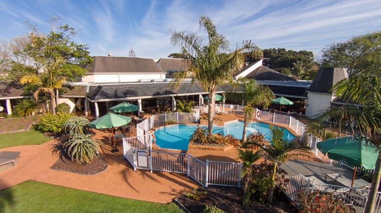 A lush subtropical hotel courtyard featuring a large outdoor swimming pool surrounded by palm trees and guests relaxing on loungers.