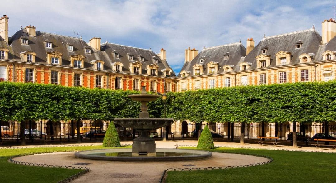 Les Jardins Du Marais 飯店寬敞的鵝卵石庭院,配有紅色遮陽傘、棕櫚樹和裝飾藝術風格的外牆。