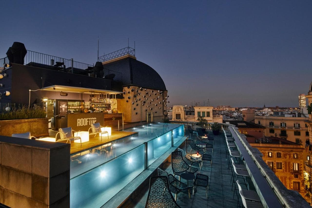 A swimmer underwater in a glass-walled rooftop pool at Ohla Barcelona, with the historic architecture of Via Laietana visible through the pool wall.