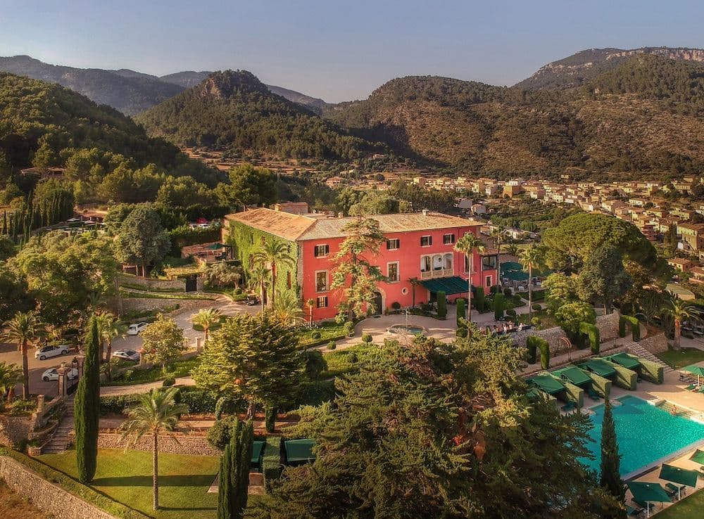 The pink stone facade of Grand Hotel Son Net viewed from the pool area, framed by palm trees and the Tramuntana mountains.