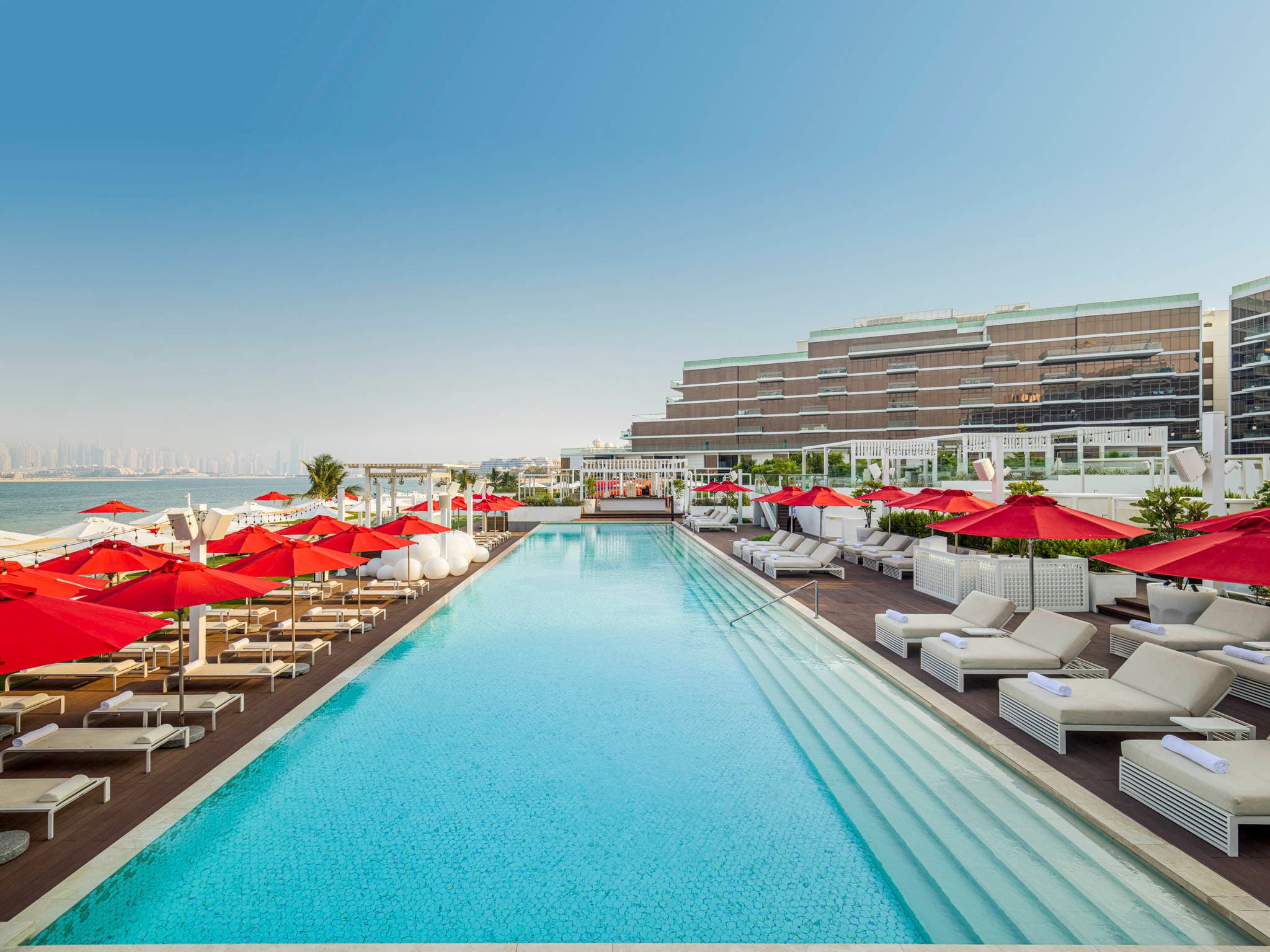 Red umbrellas and white cabanas at Fluid Beach Club, Th8 Palm Dubai, framing a view of the Dubai Marina skyline.