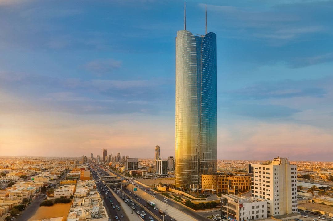 A low-angle shot of the JW Marriott Riyadh's modern facade at dusk, with the illuminated Burj Rafal tower rising against a deep blue sky.