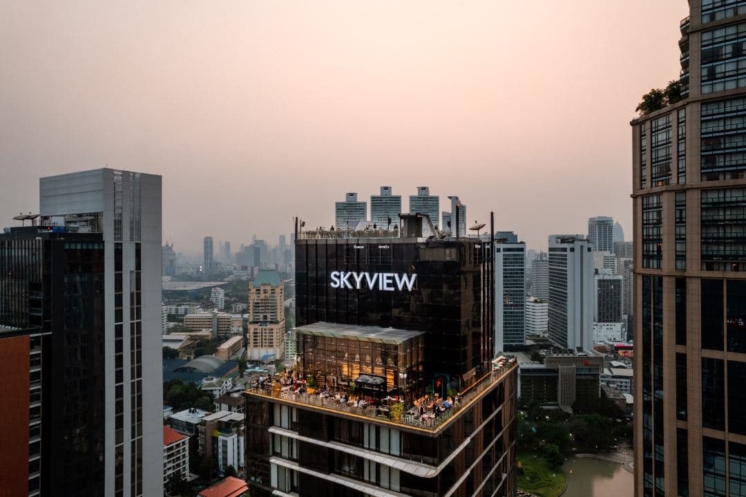 해질 무렵, SKYVIEW 호텔 인피니티 풀의 유리 난간에 기대어 있는 붉은 드레스의 여성. 배경으로 방콕 스카이라인과 엠디스트릭트의 고층 빌딩이 보인다.
