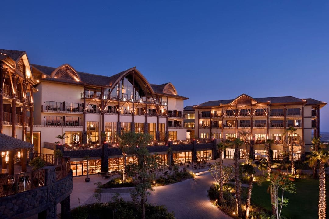 A lush, Polynesian-themed courtyard at Lapita Hotel Dubai with a winding lazy river and thatched-roof structures at sunset.