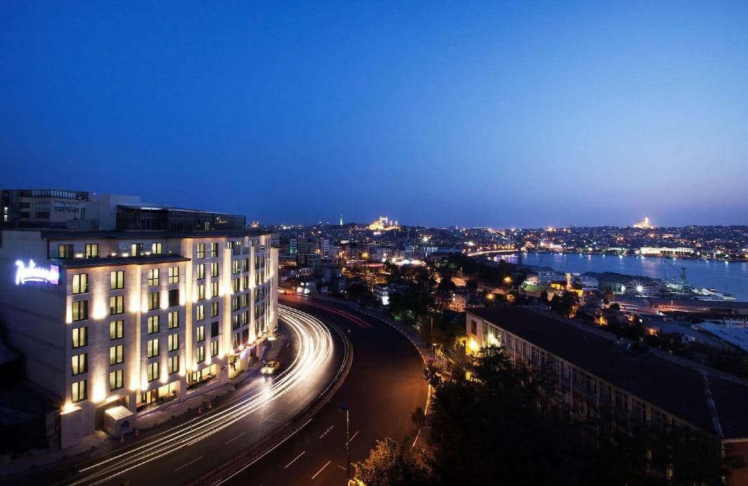 Rooftop terrace of Radisson Blu Istanbul Pera featuring Hamdi Restaurant tables set with white linens, overlooking the Golden Horn and Istanbul historic skyline at sunset.