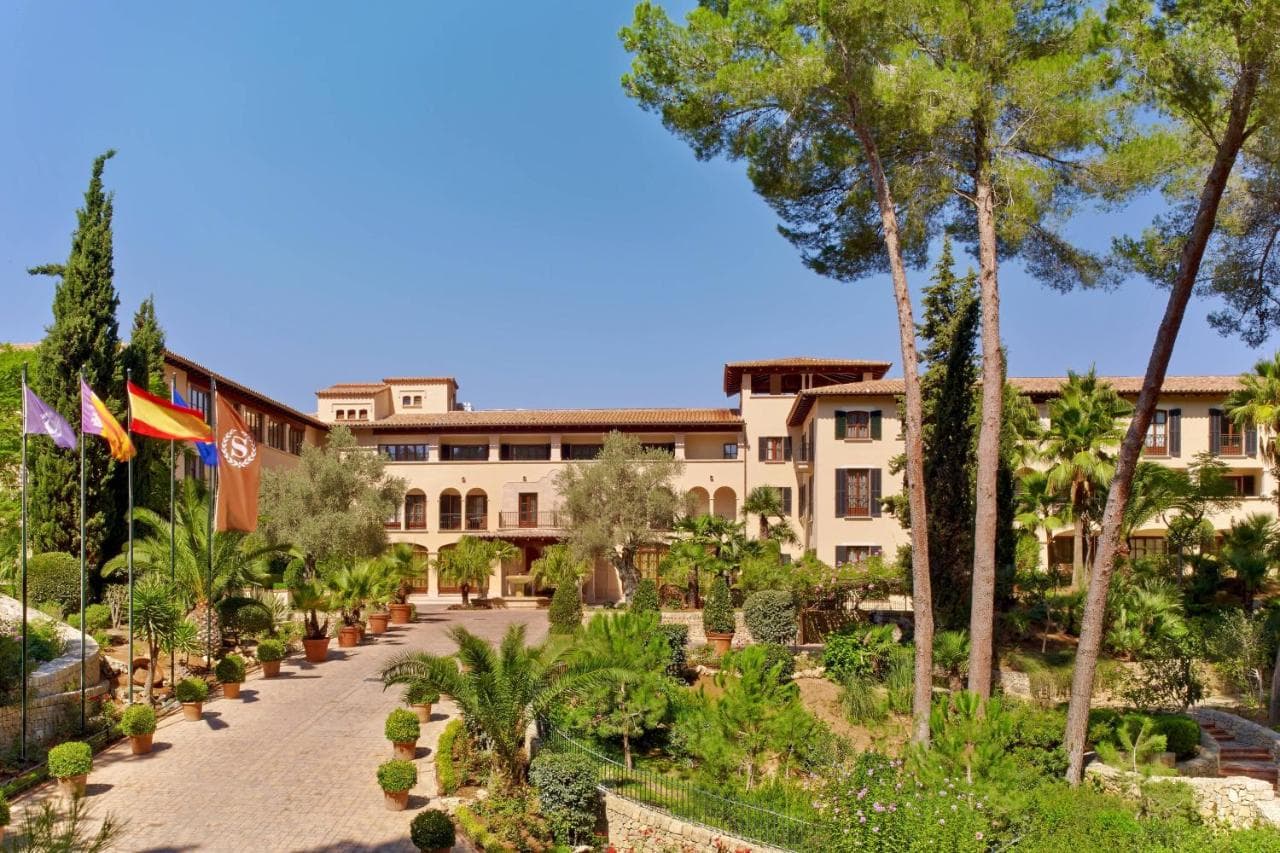A terracotta-tiled Spanish manor hotel surrounded by lush green golf courses and Mediterranean gardens, featuring a pool area with lounge chairs.