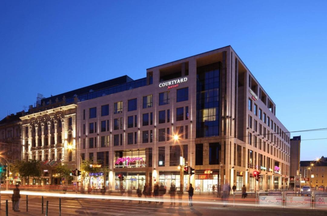 Modern hotel facade of Courtyard Marriott Budapest at twilight, illuminated against the bustling Blaha Lujza Square traffic.