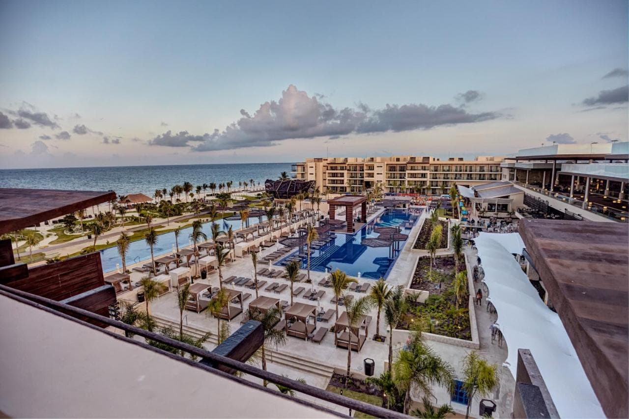A sleek, modern infinity pool at Royalton Riviera Cancun at twilight, reflecting palm trees with the illuminated Sky Lobby terrace in the background.