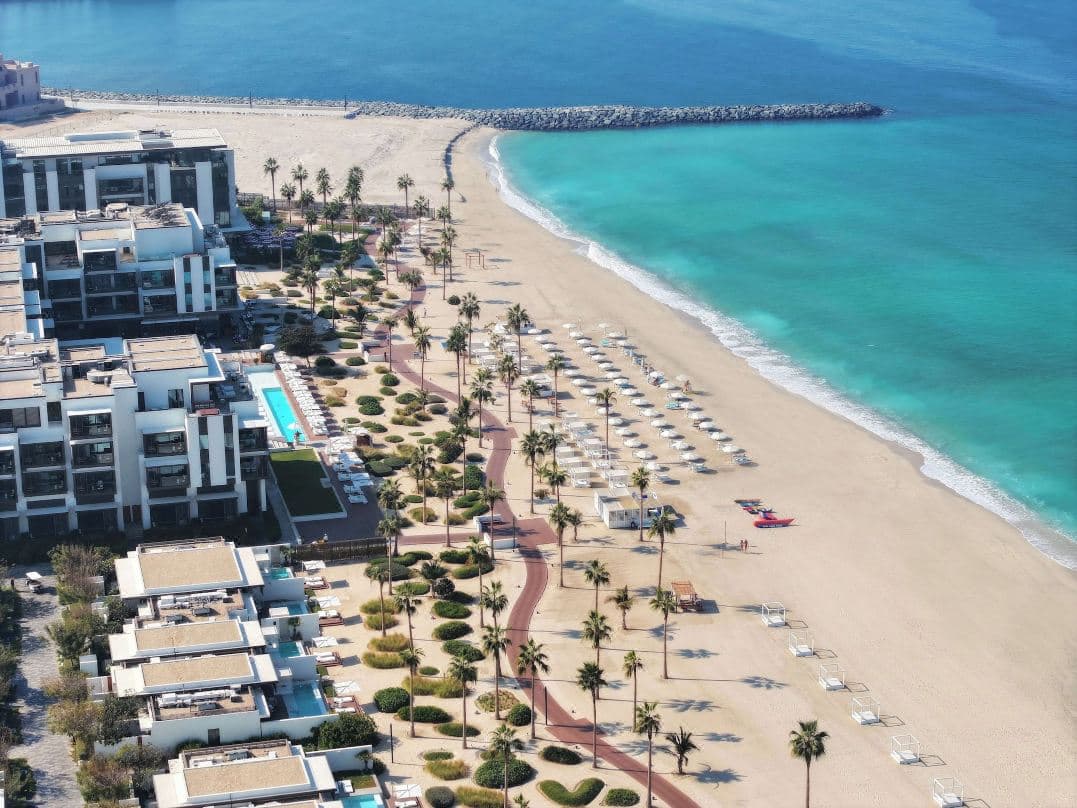 A sleek, all-white pool deck at Nikki Beach Resort & Spa Dubai featuring white tepee cabanas, turquoise water, and a giant dreamcatcher swing against a blue sky.