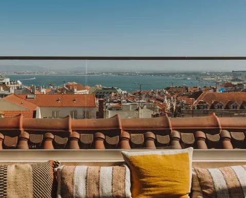 Lumi Rooftop terrace at sunset featuring the 'El Gringo' cocktail, wicker chairs, and a panoramic view of St. George's Castle and the Tagus River.