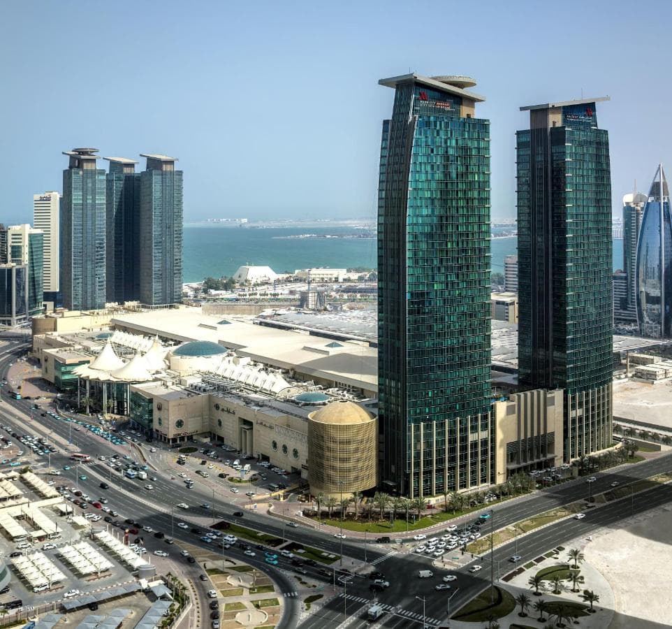 A dramatic low-angle shot of the Marriott Marquis twin towers in Doha piercing the blue sky, reflecting the surrounding West Bay glass architecture.