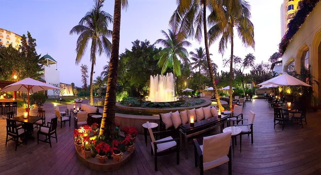 The Leela Mumbai's lush outdoor pool area featuring a cascading rock waterfall and tropical landscaping, illuminated by warm evening mood lighting.