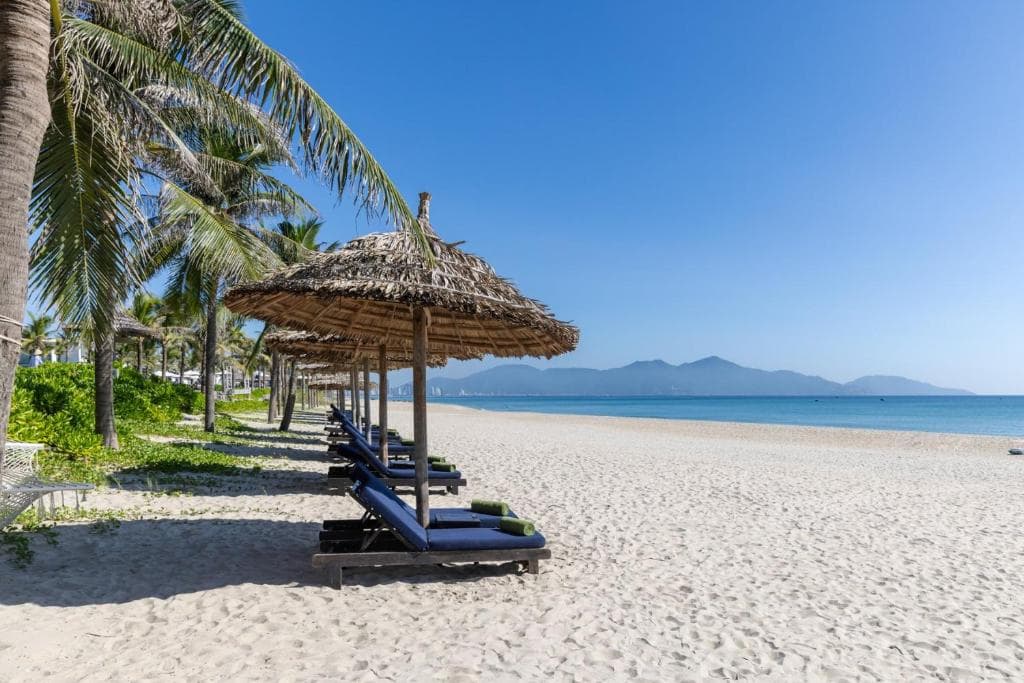 A serene sunrise view over the Meliá Danang infinity pool, with the silhouette of the Marble Mountains in the background and palm trees reflecting on the water.