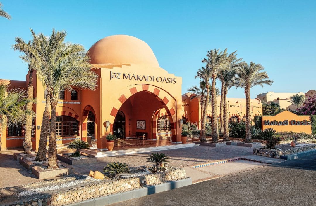 Sunset view of Jaz Makadi Oasis Resort showing traditional Nubian architecture, beige domes, and the arched bridge over the central blue lagoon pool.
