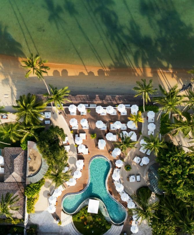 Aerial view of Nikki Beach Koh Samui showing the signature peanut-shaped Beach Club pool with teepee cabanas next to the long linear residents' pool.