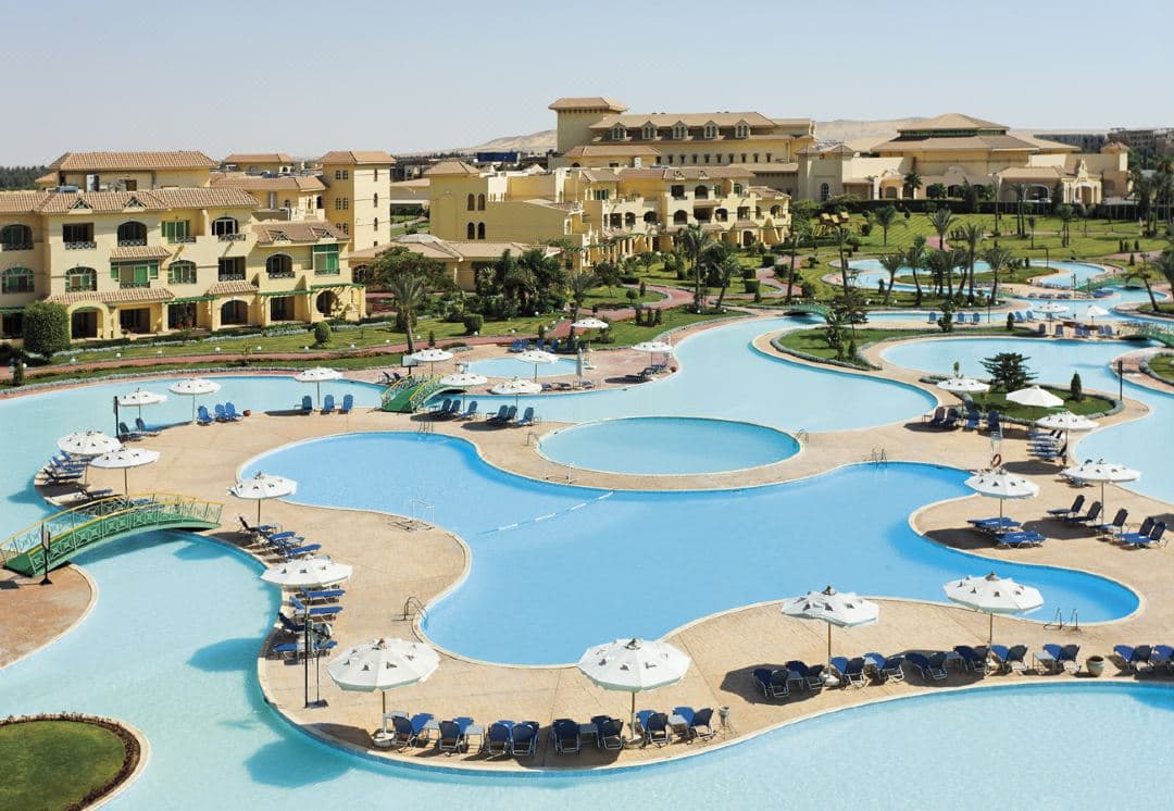 A wide shot of the Mövenpick Media City's central lagoon pool at dusk, surrounded by illuminated palm trees and low-rise terracotta buildings.