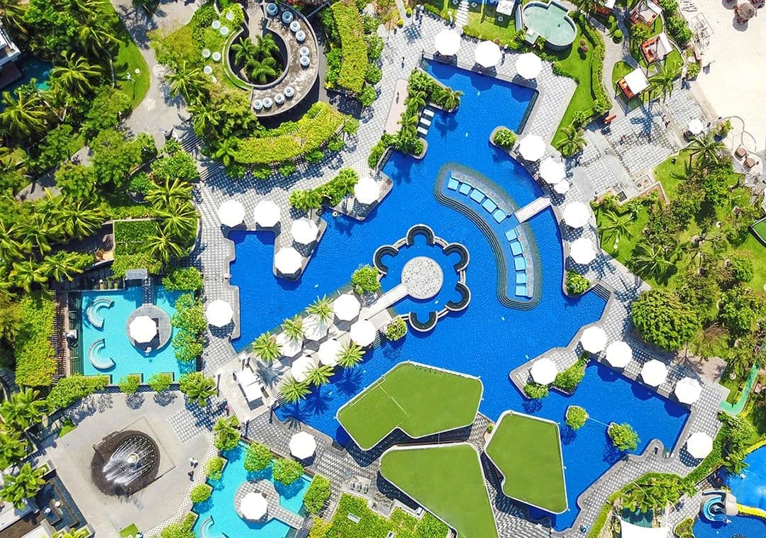 A vibrant, tropical pool complex at MGM Grand Sanya with palm trees, lounge chairs, and families playing in the water under a sunny sky.