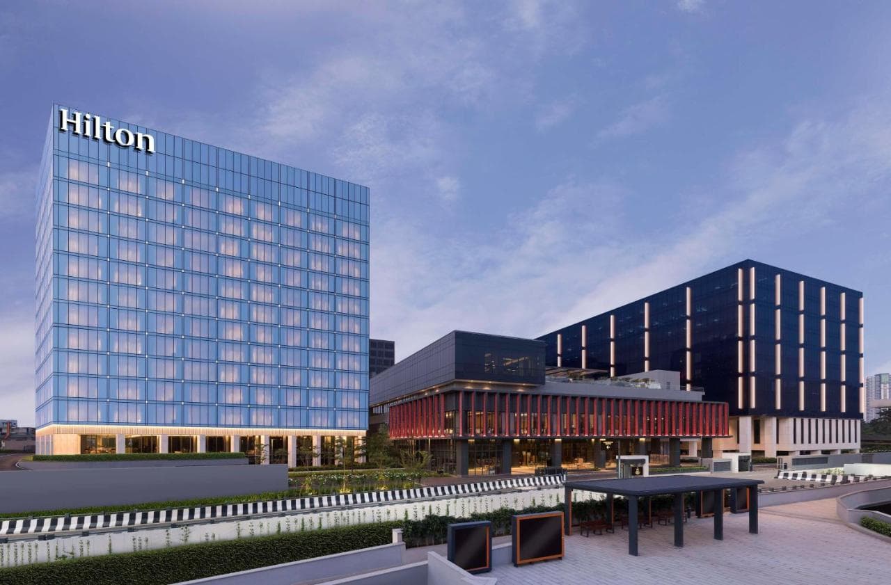 The illuminated glass facade of Hilton Bengaluru Embassy Manyata at twilight, reflecting the tech park lights with the rooftop pool glowing blue.