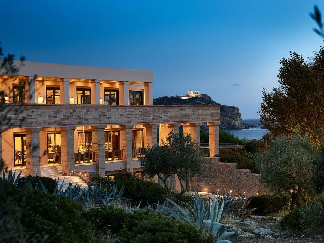 A saltwater infinity pool at Cape Sounio Grecotel with the illuminated Temple of Poseidon in the distant background at twilight.