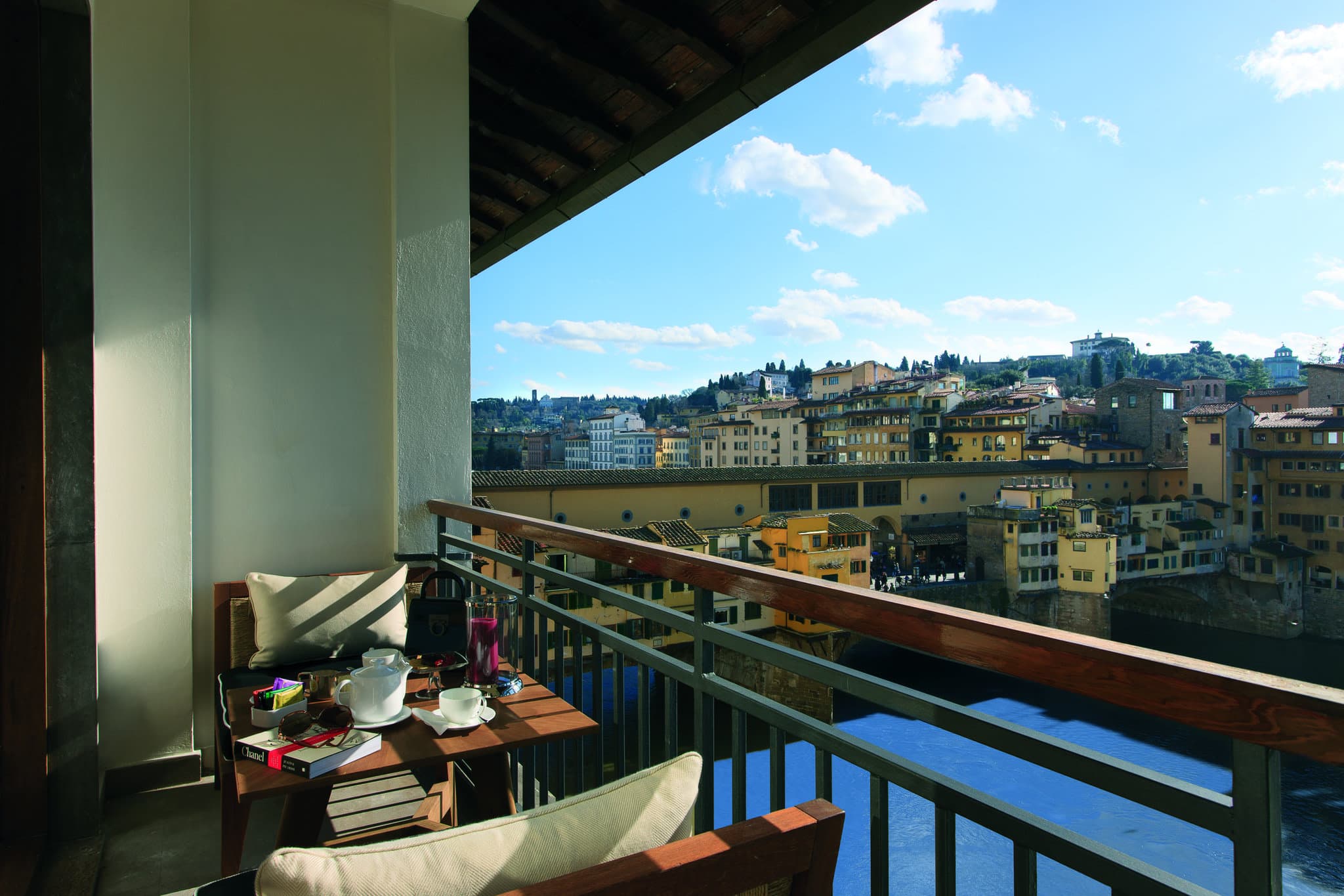View of the Ponte Vecchio and Arno River framed by the floor-to-ceiling window of a suite at Portrait Firenze, featuring mid-century modern furniture.