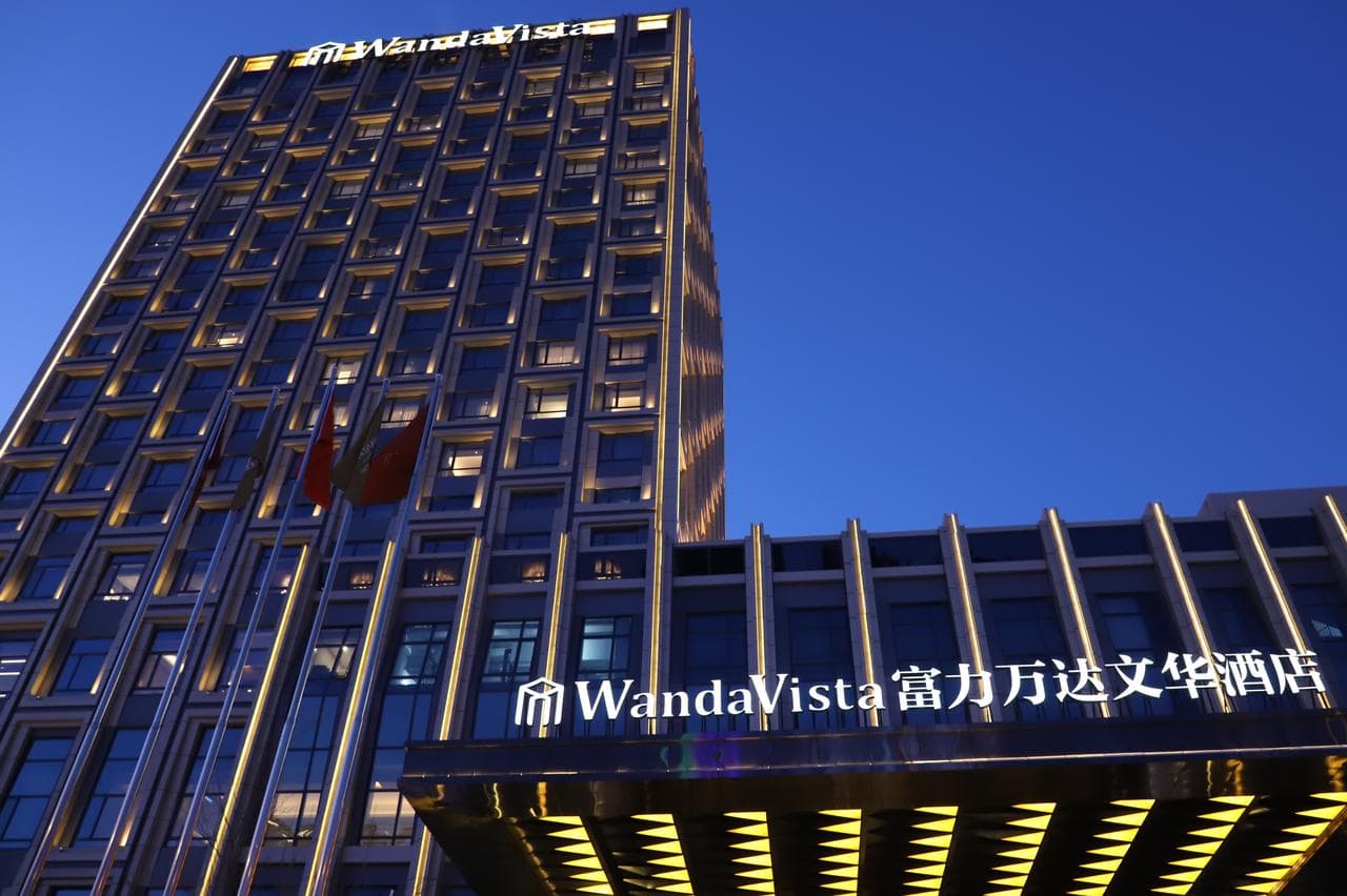 A low-angle shot of the Wanda Vista Changchun exterior at twilight, towering over the Hongqi Street district with warm light spilling from the lobby glass.