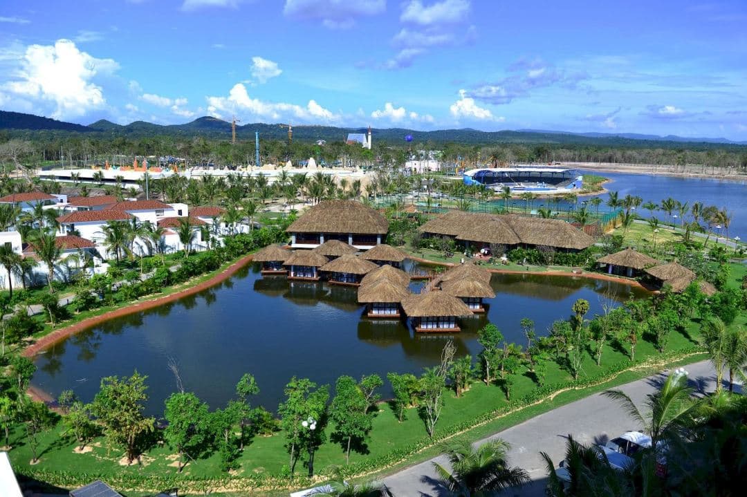 A high-angle view of the massive meandering lagoon pool at Sheraton Phu Quoc, framed by neoclassical white columns and tropical palm trees under a sunset sky.