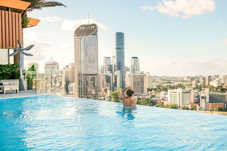 A stunning 23-meter rooftop infinity pool at Emporium Hotel South Bank at dusk, overlooking the Brisbane River and illuminated city skyline.